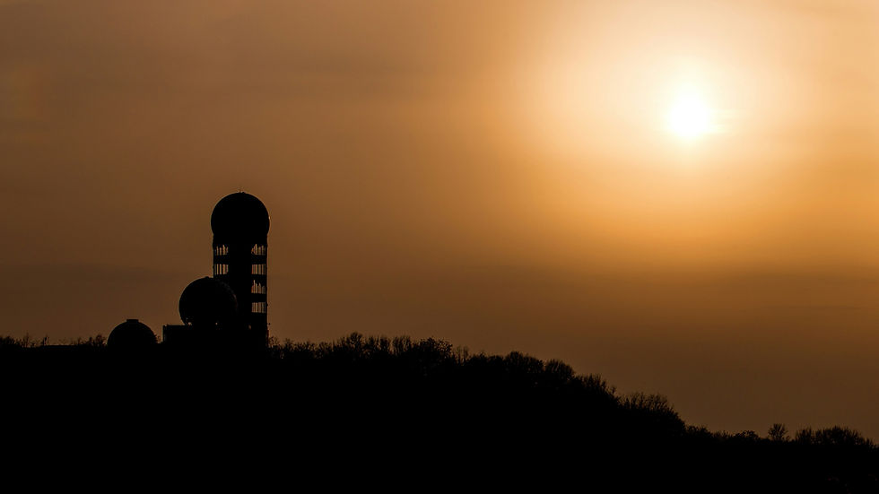 Silhouetted tower on a hill at sunset, with a glowing orange sky and bright sun overhead.