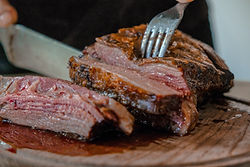 Pedaço de carne assada, sendo segurada por um garfo e fatiada por uma faca de churrasco. Está sob uma tabua de madeira, com uma pessoa usando avental preto ao fundo.