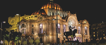 The illuminated Palacio de Bellas Artes in Mexico City stands out at night, showcasing its domed roof and ornate architecture, with a silhouetted equestrian statue in the foreground.
