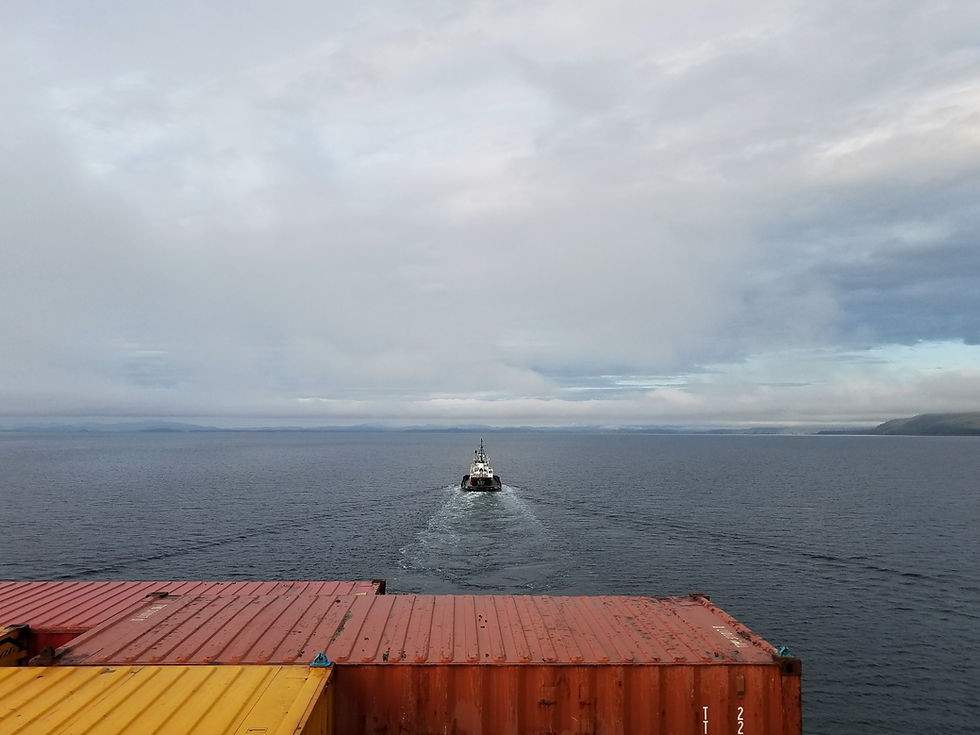 A lone tugboat sails across calm waters, trailing behind a vibrant stack of colorful shipping containers under a cloud-filled sky.