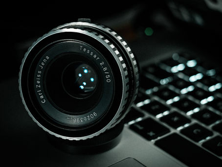 Close-up of a camera lens labeled "Carl Zeiss Jena Tessar 2.8/50" on a laptop keyboard. Dark, moody lighting with soft reflections.