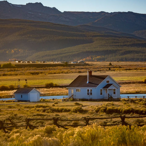 A white house and shed sit in a golden field, with mountains in the background under a clear blue sky. Rustic fence in the foreground.