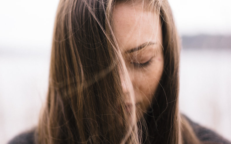 Close-up portrait of a woman with long brown hair falling over her face, eyes closed and head slightly bowed, conveying sadness
