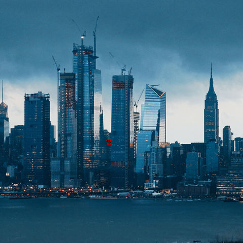 Dark skyline of a city with tall skyscrapers under a cloudy sky, lights glowing in buildings below, creating an urban evening mood.
