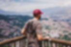Mann mit roter Kappe und grauem T-Shirt blickt von einem Geländer auf eine Stadtlandschaft mit Bergen. Klarer Himmel, entspannte Stimmung.
