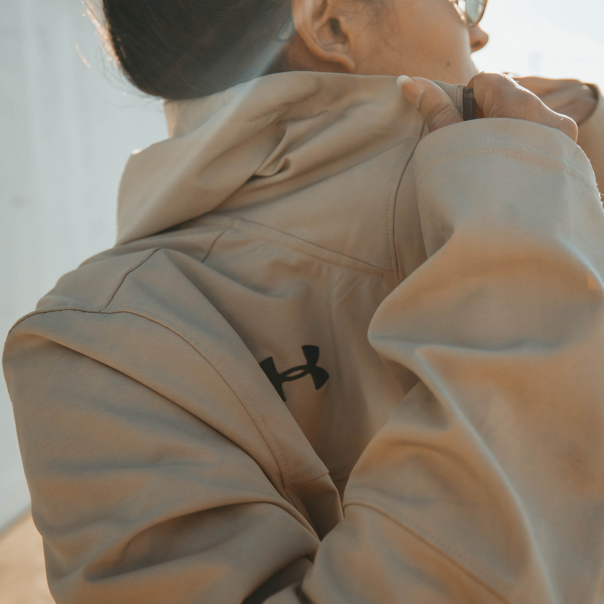 Backlit woman in sunglasses adjusts beige hoodie collar outdoors, with Under Armour logo visible at Dubai Frame tourism attraction.