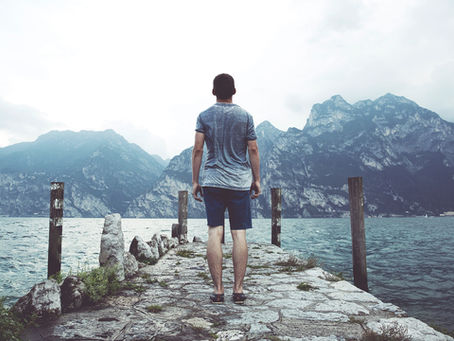 Man in shorts and t-shirt stands on a rocky pier, facing misty mountains across a lake. Overcast sky creates a calm mood.