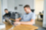 Smiling man working on a laptop in a modern office, with a colleague in the background