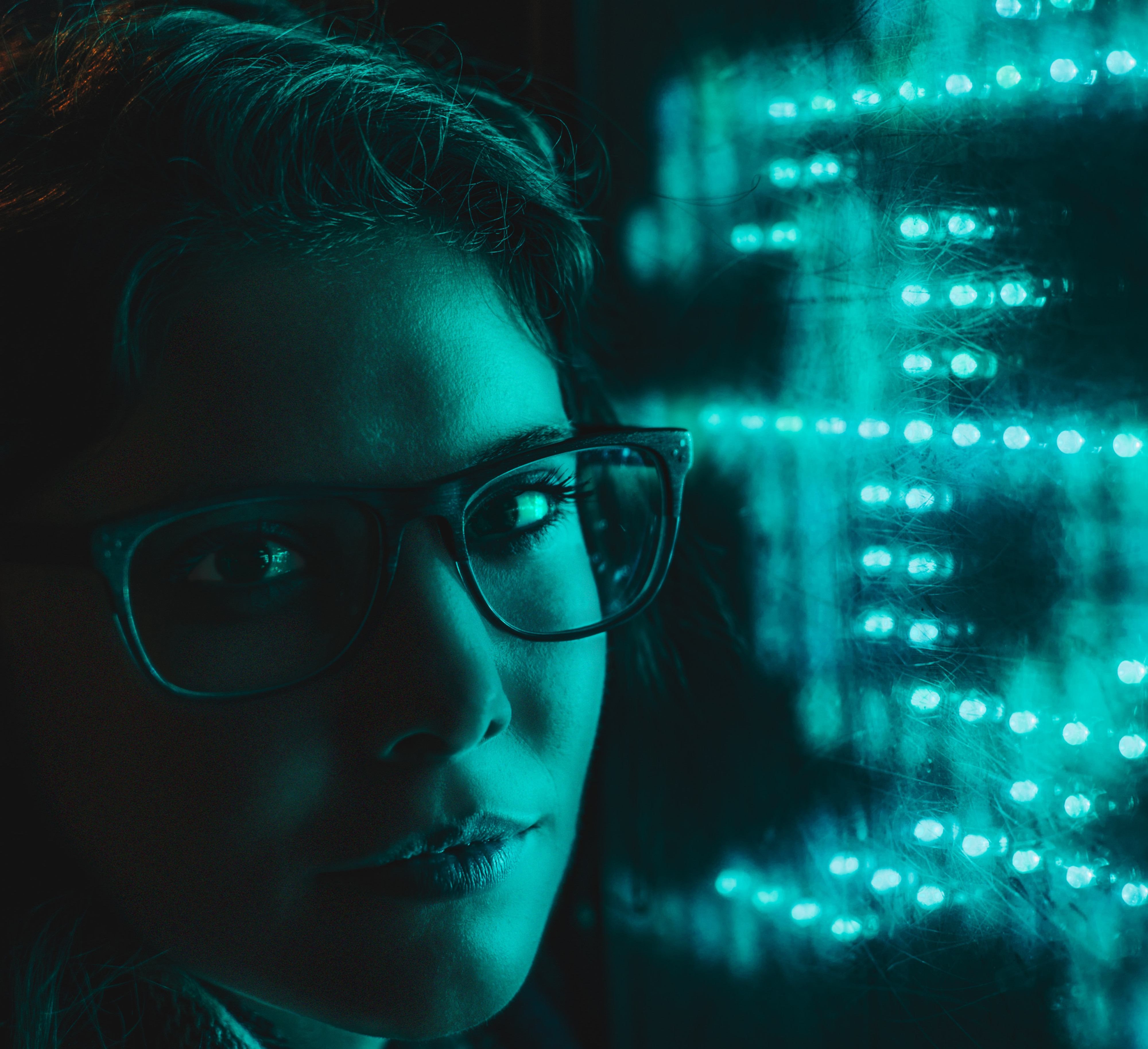 Close-up of a woman wearing glasses, lit by teal glow, gazing at bright LED lights in a dark, moody setting.
