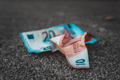 Crumpled 10 and 20 Euro banknotes lie on a textured, gray pavement. The colors are vibrant, contrasting with the muted background.