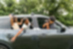 Children leaning out of a car window laughing as an adult takes their photo. Car is parked in a grassy area with trees in the background.