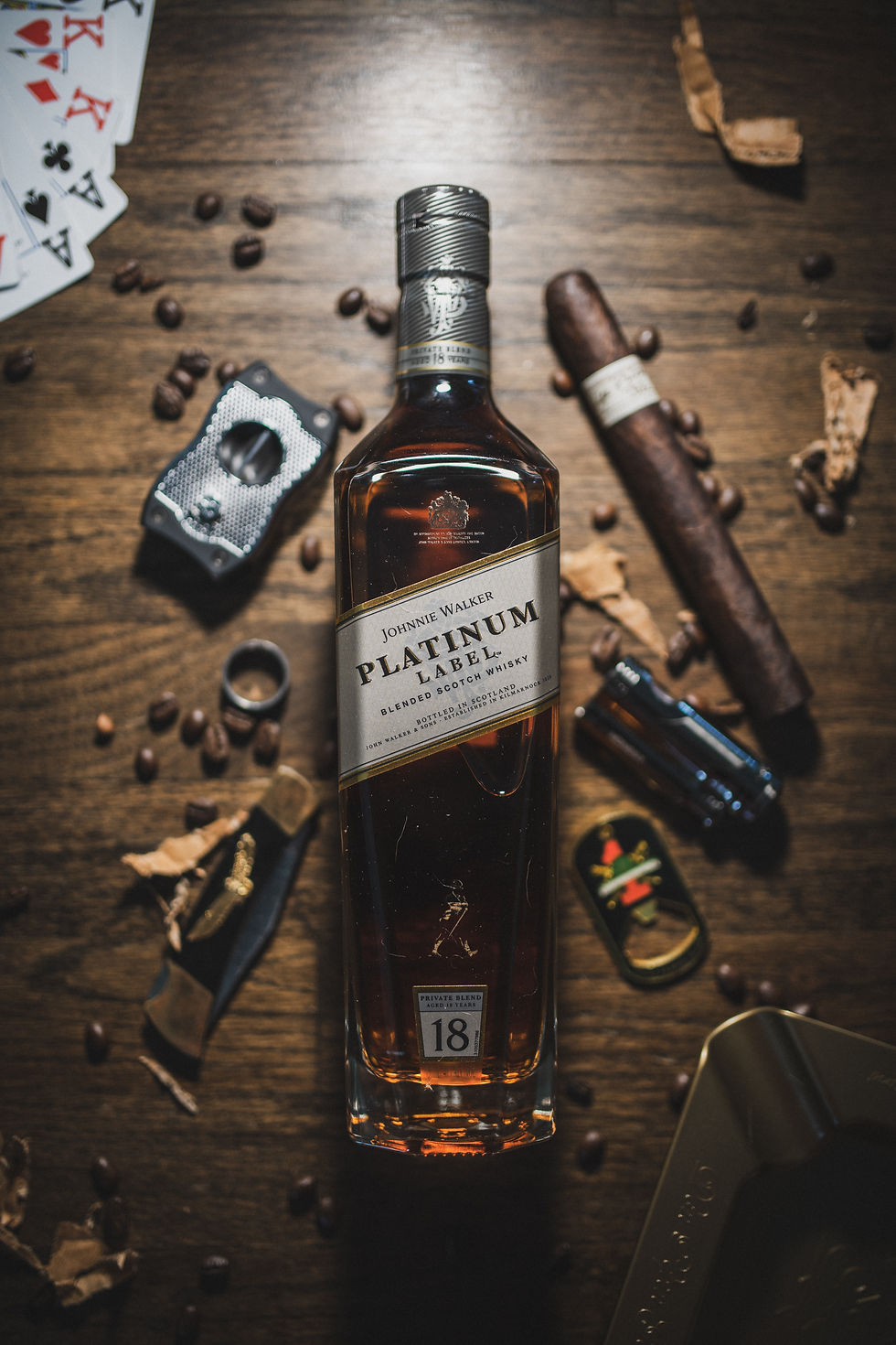 Alcohol bottle on a wooden table next to a deck of cards circled by a cigar, cigar cutter, folding knife, and lighter