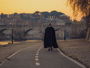 Man in black cloak walks on pathway toward stone bridge at sunset. Cobblestone path, vintage cityscape, and soft orange sky in background.