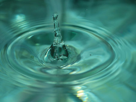 Close-up of a water droplet splashing, creating ripples in an aqua-colored liquid. The drop is rising against a blurred background.