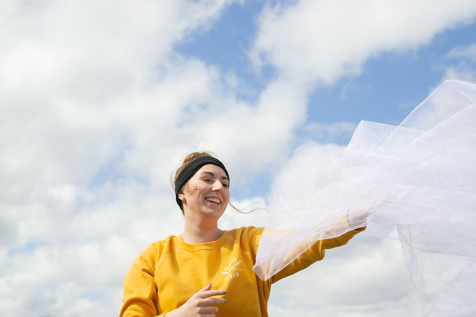 Young woman in yellow holding white fabric smiling against a cloudy sky