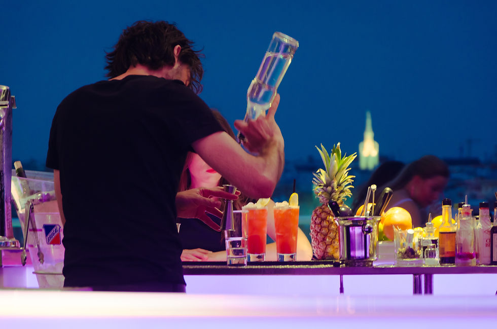 Bartender pours a drink at a colorful rooftop bar with pineapple and cocktails, blue evening skyline in the background.
