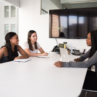Three women sit at a white conference table, engaging in conversation. A laptop and notebook are on the table. A large screen is visible.