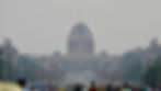 Wide boulevard leading to a large domed building, Rashtrapati Bhawan, flanked by street lamps and trees. People and vehicles visible, overcast sky.