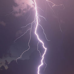 Bright lightning bolt strikes against a dark, cloudy sky. Purple hue surrounds, creating a dramatic, intense atmosphere over a distant landscape.
