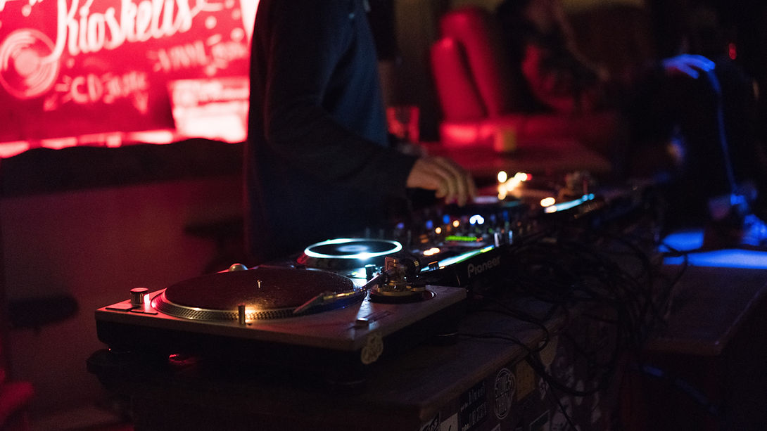 DJ stands at a turntable setup in a dimly lit club with red neon lights