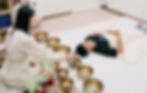 Woman in white conducts sound healing with Tibetan bowls around a lying person covered with a blanket in a serene room setting.