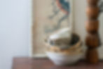 Stacked ceramic bowls on a wooden table beside a carved wooden candlestick, with a framed bird print in a calm, rustic room. Wabi sabi.