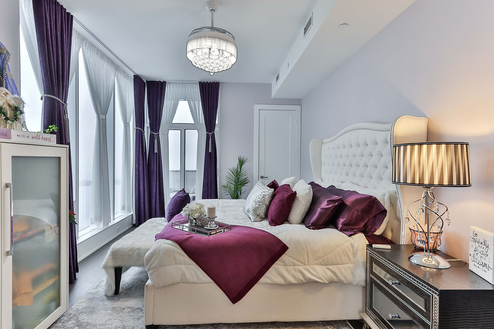 Luxurious bedroom with a white bed, purple pillows, and a furry bench. Chandelier above, purple curtains, striped lamp, and decorative tray.