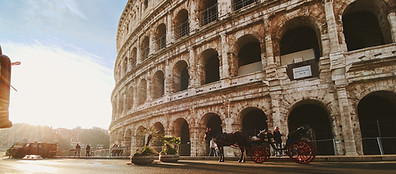 The photo shows the exterior of the Colosseum in Rome, Italy, with sunlight streaming in from the left and a few people walking nearby on the ancient stone pavement.