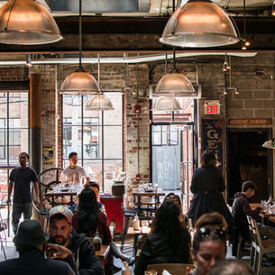 Group dining outdoors at a long table with drinks and food, surrounded by wooden beams and lights. Lively, social atmosphere.
