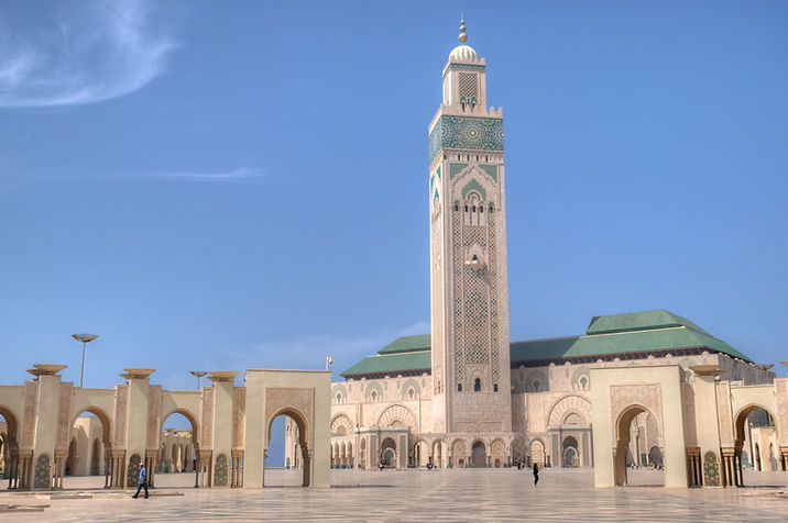 Hassan II Mosque in Casablanca, Morocco, with its towering minaret, the tallest in the world rising above intricate Moroccan architecture. The mosque sits beside the Atlantic Ocean, framed by a vast open plaza and detailed tilework under a clear sky.