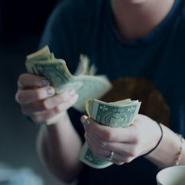 Person counting a stack of US dollar bills at a table, with a mug nearby, in a dim indoor setting.