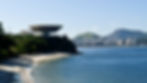 Futuristic building on a cliff by the ocean in Niteroi, with a sandy beach below. Distant city skyline and green hills under a clear blue sky.