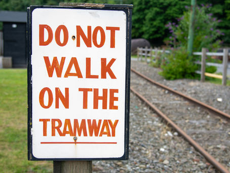 Sign reads "DO NOT WALK ON THE TRAMWAY" in bold red letters, set near railway tracks with green foliage in the background.