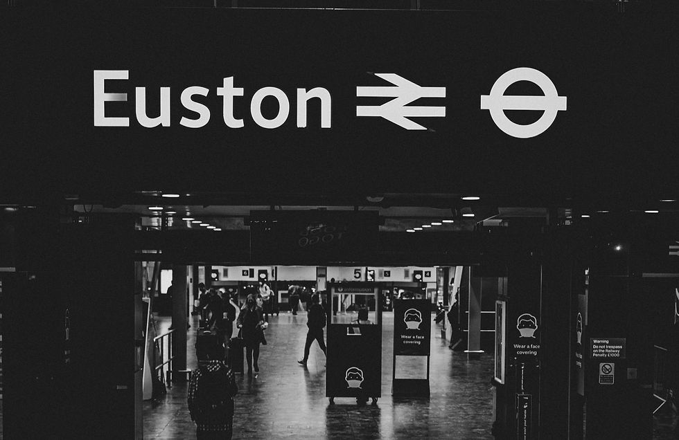 Black and white image of Euston station entrance. People walk inside, some carry luggage. Signs advise wearing face coverings. Moody ambiance.