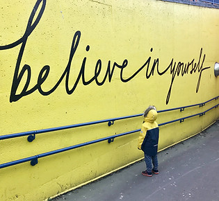 child looking up a yellow wall with black writing that says "believe in yourself"