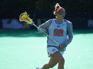Lacrosse player in uniform with number 22, wearing goggles, holds stick with a yellow ball on a green field. Bright sunlight, focused expression.
