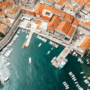 A stunning aerial view of Dubrovnik, Croatia, showcases its historic harbour with boats docked near the iconic city walls and vibrant red-tiled roofs, reflecting the charm and heritage of the medieval old town.