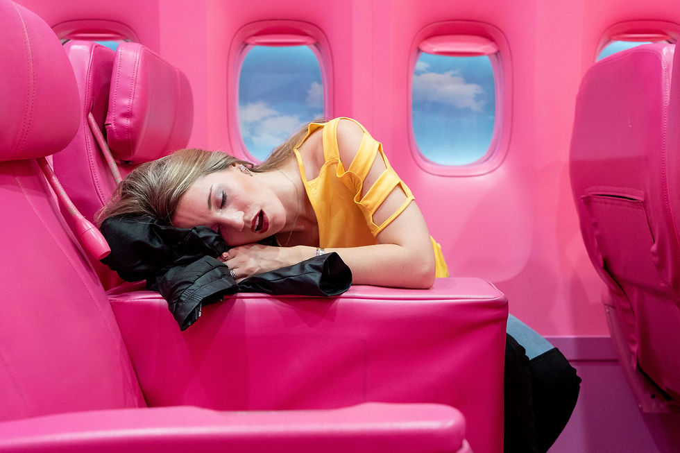 Woman in yellow top sleeps on pink airplane seat, resting on a jacket. Background displays blue sky through windows, creating a dreamy mood.