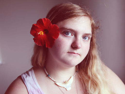 Ung, autistisk kvinna med långt hår och röd blomma bakom örat. Hon bär halsband och ett vitt linne, ser neutral ut mot en ljus bakgrund.