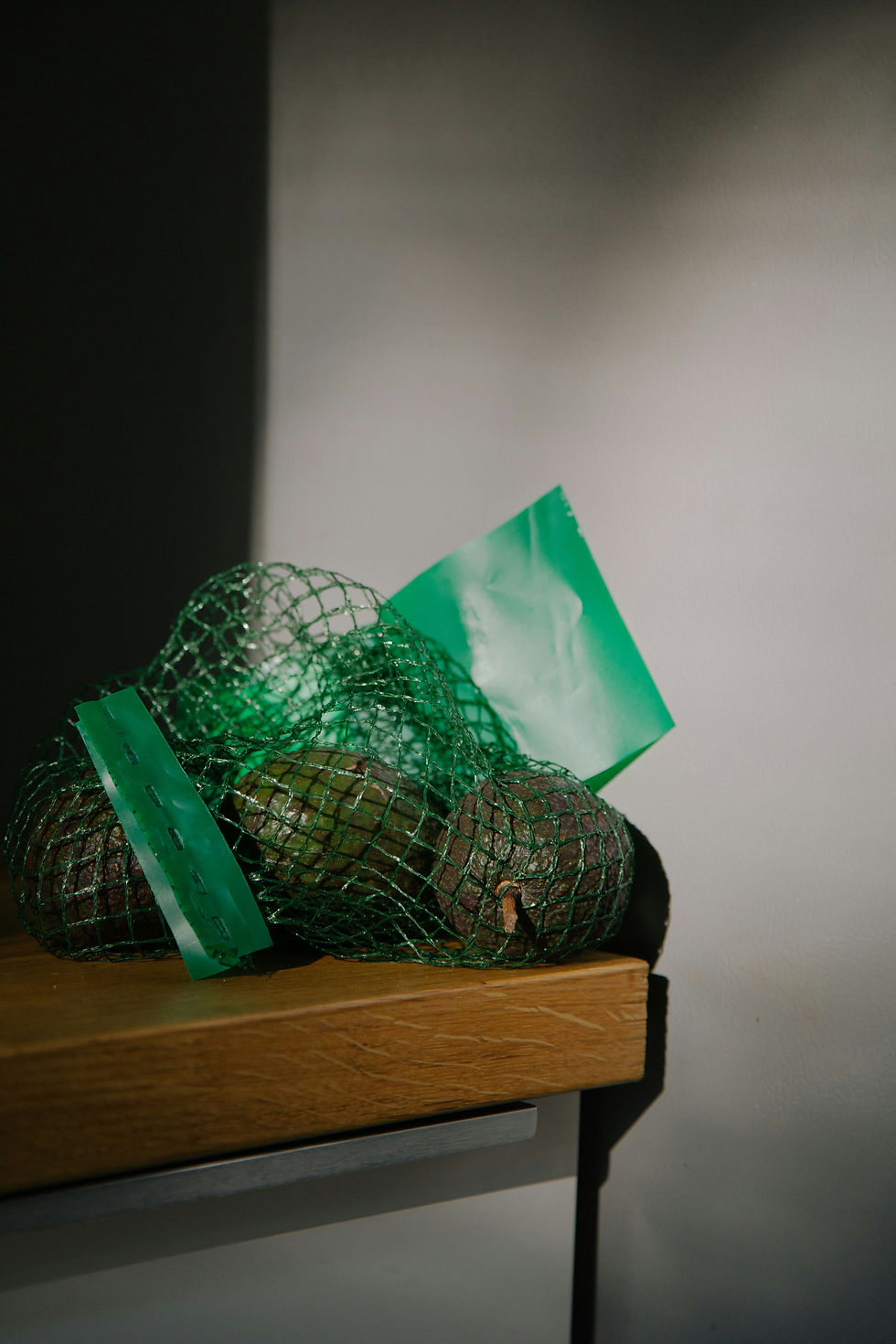 Avocados in a green mesh bag sit on a wooden surface, with soft light casting shadows on the neutral background.