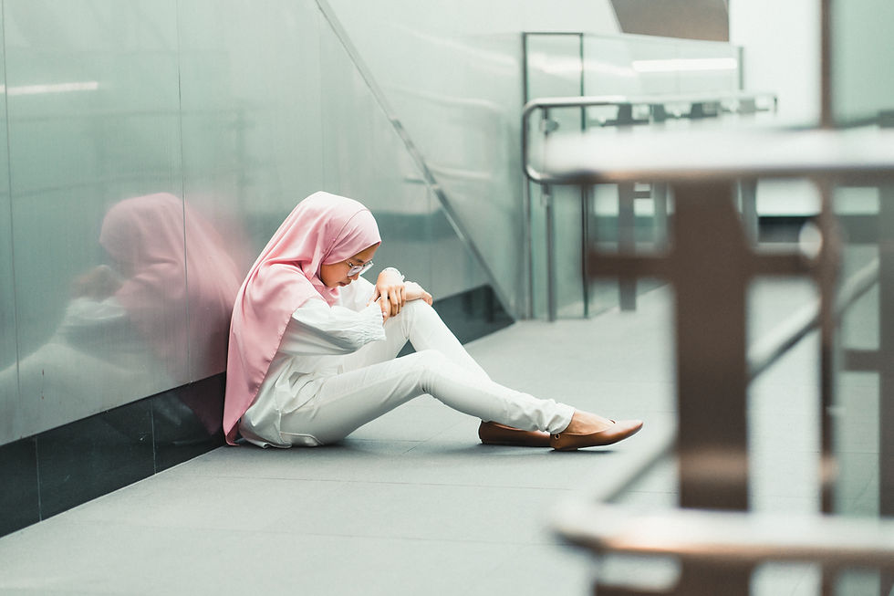 En kvinna med vvita kläder och rosa hidjab sitter på golvet och är nedstämd