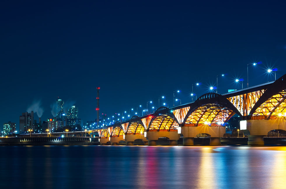 Illuminated bridge arches over a calm river at night, with city skyline in the background. Bright streetlights and reflections create a serene mood.
