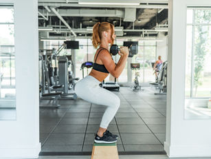 Woman in gym doing a squat on a box while holding dumbbells. She's wearing black and white activewear. Bright, modern gym setting.