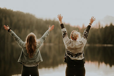 Image of two women with their hands raised high shouting out over a lake that is surrounded by woodland