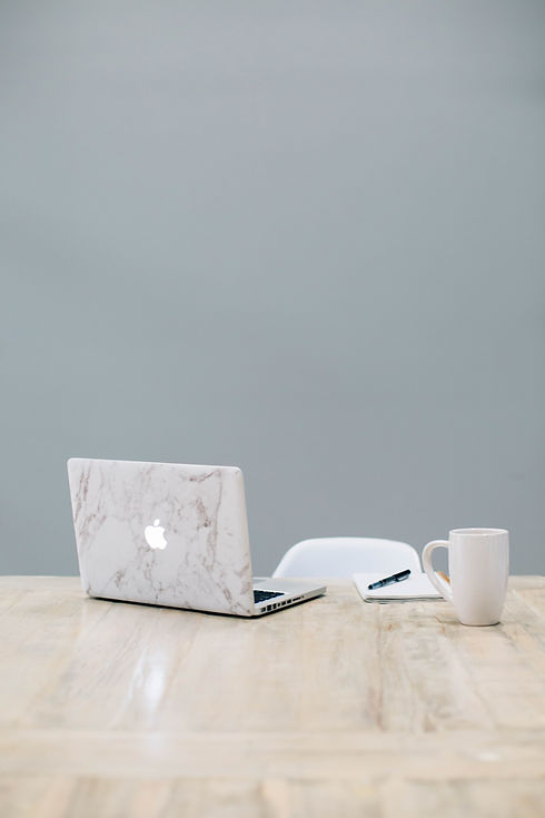 Picture of a macbook, notepad, pen and mug on a empty desk. Top of white desk chair can be seen