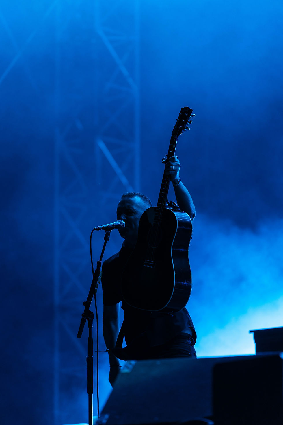 Musician on stage under blue lights, holding a guitar aloft by microphone. Silhouette against a misty backdrop, conveying energetic mood.