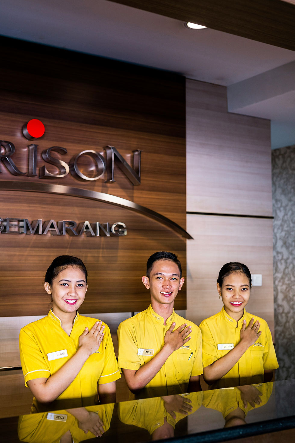Three smiling hotel staff in yellow uniforms stand with hands on their chests. A wooden wall with partial signage is behind them.