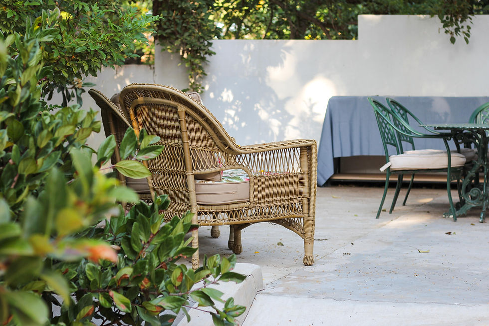 Wicker chairs and a table with a blue cover on a patio, surrounded by lush green plants. Sunlight casts soft shadows, creating a tranquil mood.