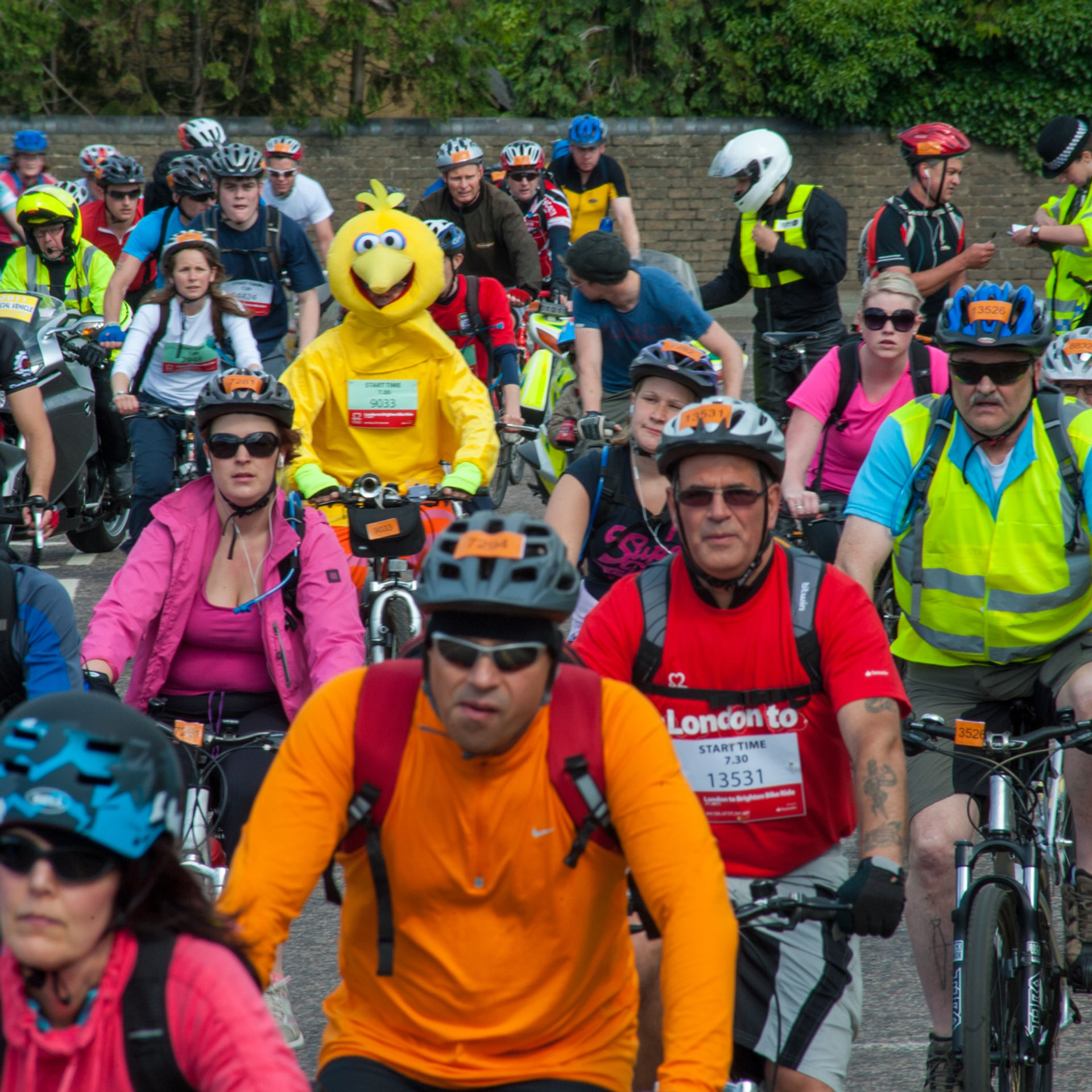 Cyclists in colorful gear, including one in a yellow bird costume, ride on a city street. Police and spectators watch from the background.