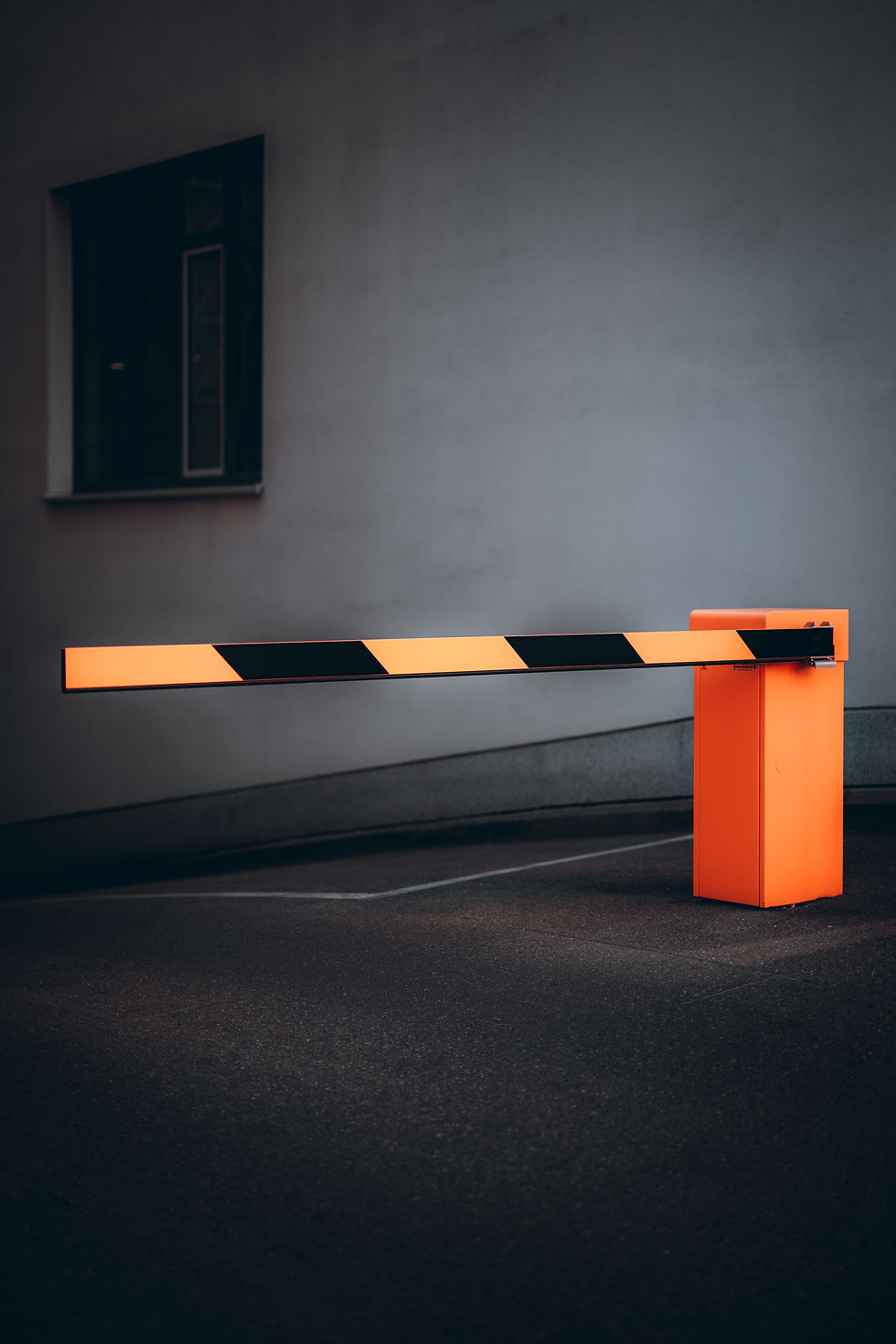 Orange and black barrier arm, set down in a dimly lit parking area; background is a plain grey wall with a window.